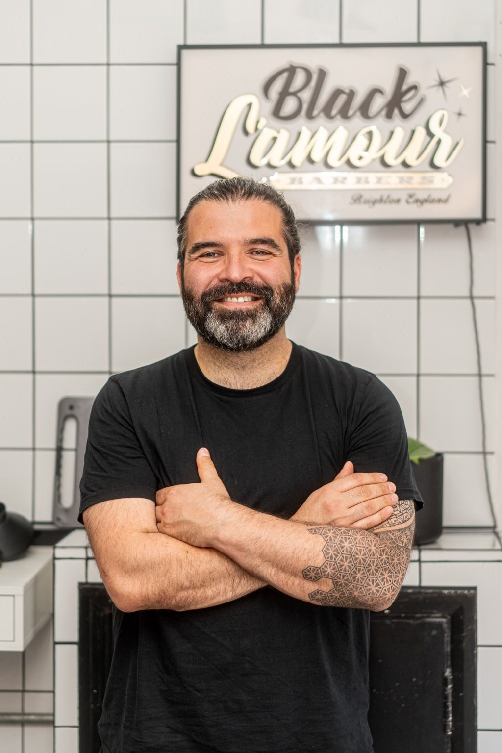 Hairdresser with tattoos and beard, arms crossed; backdrop: Black Lamour Bakery. Known for using Olivia Gardens premium tools.